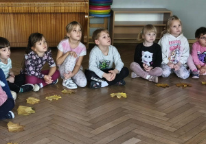Na zdjęciu widzimy grupkę dzieci, przed którymi leżą liście. Dzieci słuchają Pani, która tłumaczy, jakie mają zadanie.