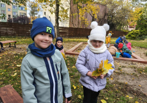 Na zdjęciu widzimy chłopca, który się uśmiecha oraz dziewczynkę, która trzyma w rękach zebrane kolorowe liście.