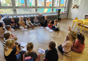 Dzieci słuchają prowadzącej siedząc w kręgu.