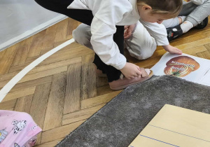 Na zdjęciu widzimy dziewczynkę, która kładzie na podłodze ilustrację przedstawiającą dynię turbanową.