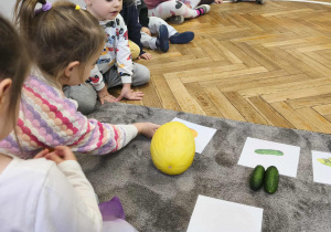 Na zdjęciu widzimy dziewczynkę, która przyporządkowuje ilustrację melona do owoca leżącego na dywaniku.