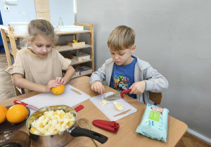 Na zdjęciu widzimy chłopca, który kroi kawałek jabłka oraz dziewczynkę, która roluje pomarańczę.