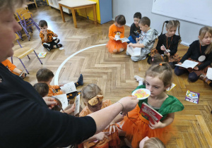Na zdjęciu widzimy grupkę dzieci, które otrzymują medal uczestnictwa w "Balu Dyni" oraz łamigówki i kolorowanki.