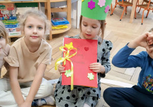 Dzieci siedzą na podłodze, a Jubilatka trzyma książeczkę z laurek dzieci.