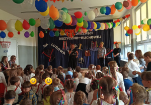 Zdjęcie przedstawia dzieci zgormadzone na sali. Dzieci słuchaja przemówienia pani dyrektor z PM 155. W tle znajduje się dekoracja.