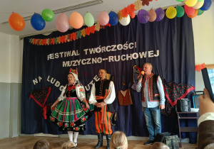 Zdjęcie przedstawia troje osób w strojach ludowych. Na drugim planie znajduje się dekoracja.