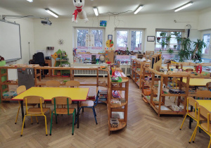 na zdjęciu znajduje się sala grupy czwartej, niskie stoliki i regały, na których znajdują pomoce rozwojowe dla dzieci, żółte stoliki, po lewej stronie na ścianie wisi tablica multimedialna