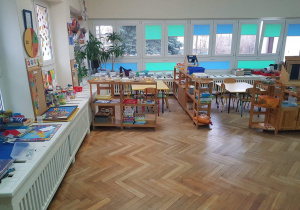 na zdjęciu znajduje się sala grupy czwartej, niskie stoliki i regały, na których znajdują pomoce rozwojowe dla dzieci, widać okna z niebieskimi i zielonymi żaluzjami