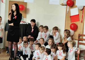 Pani stoi i przemawia do mikrofonu, dzieci siedzą.