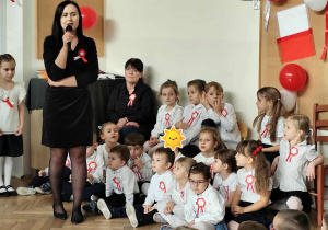 Dzieci siedzą, apani przemawia przez mikrofon.