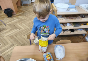 Na zdjęciu widzimy chłopca, który odmierza za pomocą szklanki odpowiednią ilość mąki.