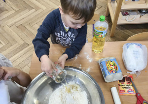 Na zdjęciu widzimy chłopca, który wlewa odmierzoną ilość oleju do miski z pozostałymi składnikami.