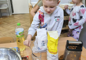 Na zdjęciu widzimy chłopca, który odmierza odpowiednią ilość cukru za pomocą szklanki.