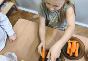 Na zdjęciu widzimy dziewczynkę, która trze marchewkę na tarce o grubych oczkach.