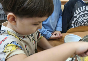 Na zdjęciu widzimy chłopca, który odmierza za pomocą szklanki odpowiednią ilość jogurtu naturalnego potrzebnego do ciasta.