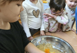 Na zdjęciu widzimy dziewczynkę, która wkłada do dużej miski marchewkę wcześniej ztarkowaną.