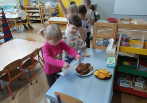 Na zdjęciu widzimy dziewczynkę, która wybiera kawałek ciasta marchewkowego, aby go zjeść. Na drugim planie widzimy dziewczynkę, która degustuje ciasta.