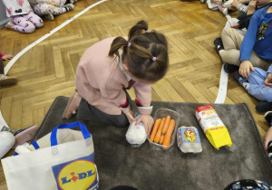 Na zdjęciu widzimy dziewczynkę, która wyjęła jeden ze składników ciasta marchewkowego z torby.