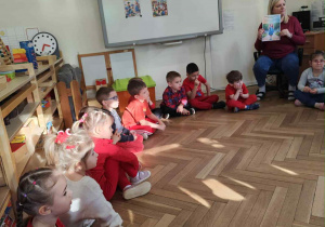 Zdjęcie przedstawia dzieci siedzące w kole podczas zajęć. Kobieta pokazuje im obrazek.