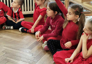 Zdjęcie przedstawia dzieci siedzące w kole. Dwie dziewczynki zgłaszają się do odpowiedzi.