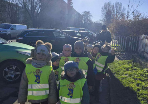 Zdjęcie przedstawia dzieci w odblaskowych kamizelkach, stojące w parach podczas spaceru.