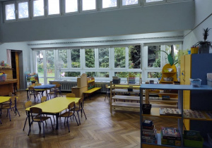 na zdjęciu widać salę przedszkolną grupy pierwszej, stoliki z żółtymi blatami, okno z widokiem na drzewa i choinki