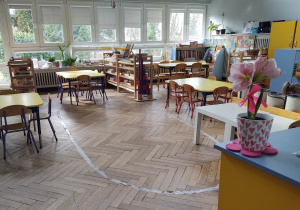 na zdjęciu widać salę przedszkolną grupy pierwszej, stoliki z żółtymi blatami, na pierwszym planie widać wyklejoną na podłodze elipsę, na dalszym planie widać niskie regały z materiałem rozwojowym dla dzieci