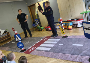 Na zdjęciu widzimy Panie ze Straży Miejskiej. Jedna z nich trzyma materiałowy sygnalizator świetlny i przypomina dzieciom, na jakim kolorze można przejść przez drogę.