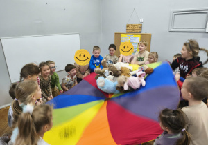 Na zdjęciu widzimy grupkę dzieci, które uczestniczą w zabawie z wykorzystaniem chusty animacyjnej. Na chuście położone są misie dzieci.