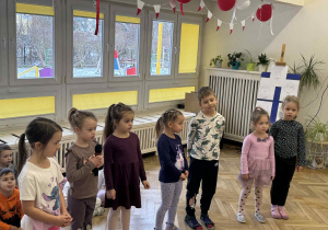 Zdjęcie przedstawia dzieci stojące w rzędzie. Jedna z dziewczynek mówi do mikrofonu.