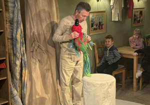 Zdjęcie przedstawia aktora na scenie, trzymającego kukiełkę papugę.