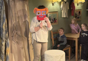 Zdjęcie przedstawia aktorkę trzymającą przy twarzy maskę, stojącą na scenie.