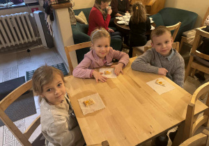 Zdjęcie przedstawia dzieci siedzące przy stolikach. Na stolikach leżą ciasteczka.