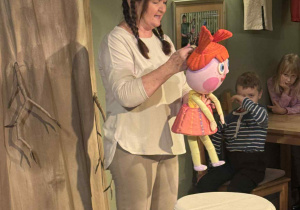 Zdjęcie przedstawia aktorkę stojącą na scenę z lalką w ręku.