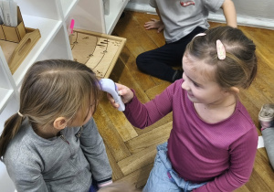 Na zdjęciu widzimy dwie dziewczynki. Jedna dziewczynka mierzy temperaturę drugiej za pomocą termometru do ciała.