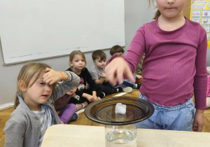 Na zdjęciu widzimy dziewczynkę, która kładzie kostę lodu na talerz. Wykonywane jest doświadczenie z deszczem w słoiku.