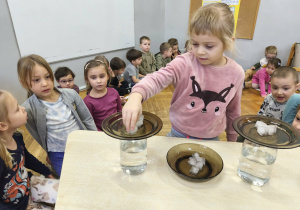 Na zdjęciu widzimy dziewczynkę, która kładzie kostę lodu na talerz. Wykonywane jest doświadczenie z deszczem w słoiku.