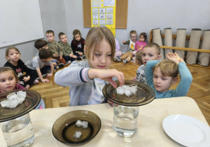 Na zdjęciu widzimy dziewczynkę, która kładzie kostę lodu na talerz. Wykonywane jest doświadczenie z deszczem w słoiku.