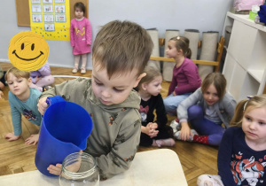 Na zdjęciu widzimy chłopca, który wlewa do słoika wodę.