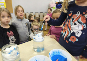 Na zdjęciu widzimy dziewczynkę, która za pomocą pipety i zabarwionej wody wlewa krople niebieskiej wody do słoika z pianką do golenia.