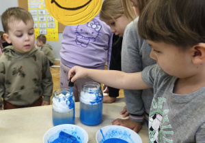 Na zdjęciu widzimy chłopca, który z wykorzystaniem pipety i niebieskiej wody tworzy w słoiku deszcz.