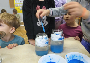 Na zdjęciu widzimy dziewczynkę, która z wykorzystaniem pipety i niebieskiej wody tworzy w słoiku deszcz.