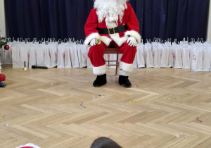 Mikołaj siedzi na krześlę, za nim widać napis mikołajki