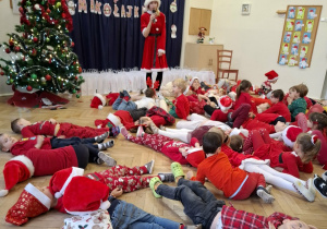 dzieci leżą na podłodze. Widać kobietę w stroju Pani Mikołajowej. Z prawej strony widać choinkę