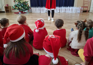 kobieta w stroju Mikołajki, stoi przed dziećmi. W tle widać napis mikołajki