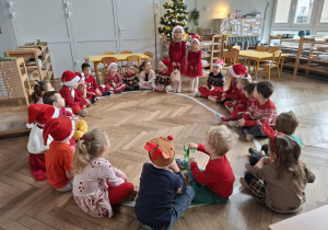 dzieci siedzą na elipsie w strojach z elementami świątecznymi. Dwie dziewczynki stoją przed nimi