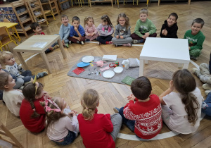 grupa siedzi na elipsie. w środku stoją dwa stoliki, na dywanie leżą elementy do ułożenia stołu wigilijnego oraz elememty, które do niego nie pasują
