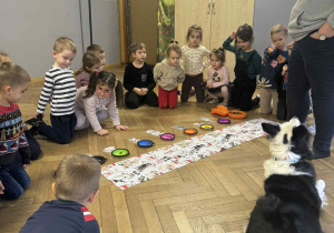 Zdjęcie przedstawia kobietę, dzieci i psa wokół kawałka materiału i kolorowych podstawków.
