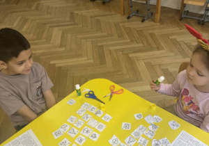 Zdjęcie przedstawia dzieci siedzące przy stoliku podczas pracy. Na stole leżą obrazki.