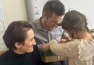 Zdjęcie przedstawia dziewczynkę rozpakowującą swój prezent, razem z rodzicami.